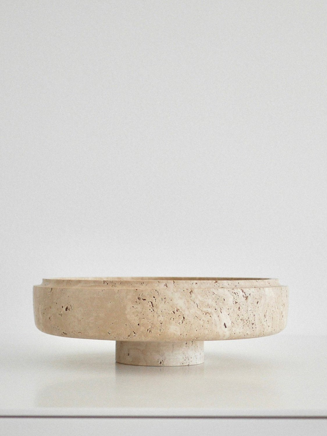 Minimalist profile shot of the Bruna Bowl in travertine, set against a neutral background, focusing on its clean lines, texture, and floating effect on the pedestal base.