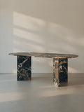 Sienae Dining Table in Dark Emperador marble with oval-edged tabletop, shown from a slight angle highlighting the V-shaped base