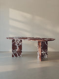 Sienae Dining Table in Rosso Levanto marble with oval-edged tabletop, shown from a slight angle highlighting the V-shaped base