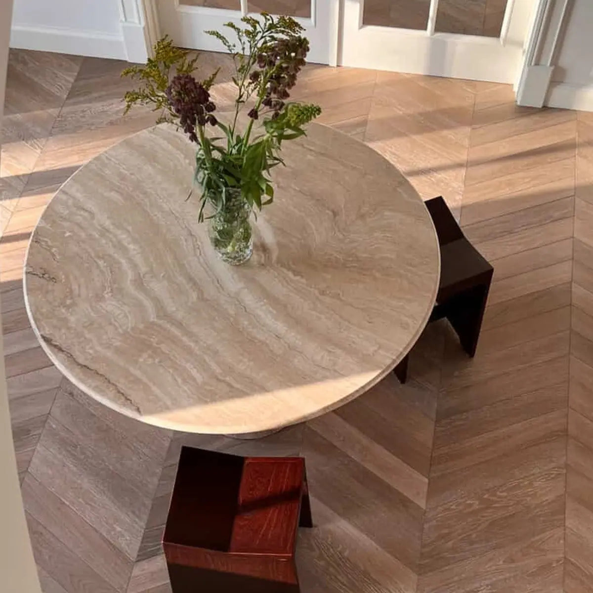 Round Leila travertine table in an interior with flowers and wooden chairs.